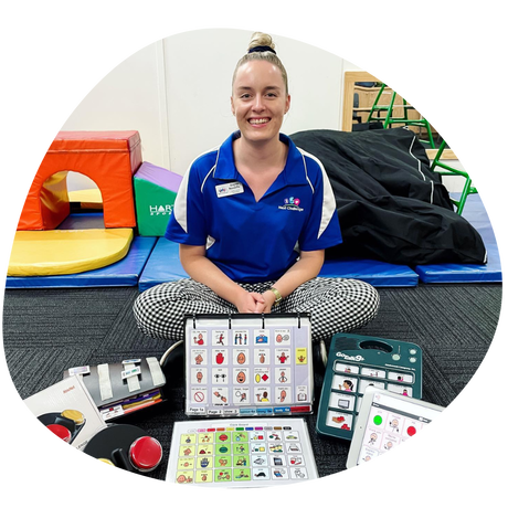 Dayna (she/her) a senior speech pathologist, wearing a Next Challenge blue polo, is sitting cross legged on the floor with various types of AAC in front of her. A range of play gym equipment and mats, a mirror, and a white wall can be seen in the background.Picture