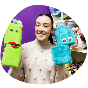ID: Speech pathologist Sienna (she/her) holds two puppets made out of milk bottles, material, and craft supplies. A purple wall and various books and therapy resources on shelves can be seen in the background. Sienna is wearing a light coloured knit with pom-poms, she is smiling and looking at the cameraPicture