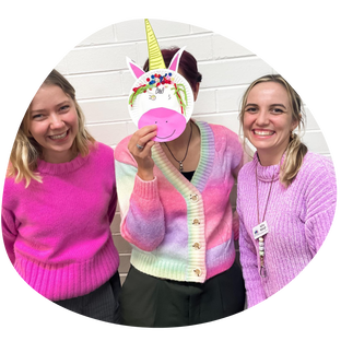 ID: Laura (left - she/her), Amy (middle - she/her) and Neli (right - she/her) from the Next Challenge OT team are standing in a row wearing pink, purple, and pastel rainbow coloured jumpers in front of a white brick wall. Amy holds a paper plate with a unicorn face in front of her face. Laura and Neli are standing on either side smiling and looking cheerfulPicture